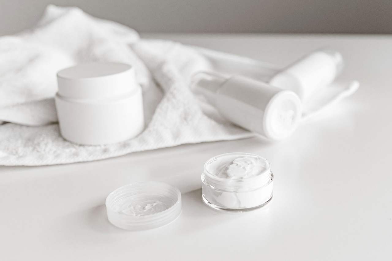 Hũ kem trắng, tuýp và lọ trơn không nhãn đặt trên khăn bông trắng, hai nắp hũ mở có kem bên trong, tông trắng sáng