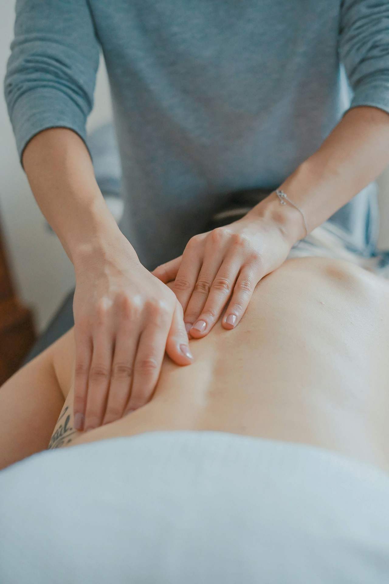 Hai bàn tay người kỹ thuật viên áo xám xanh ấn dọc lưng một người nằm sấp, khăn xanh nhạt phủ ngang hông
