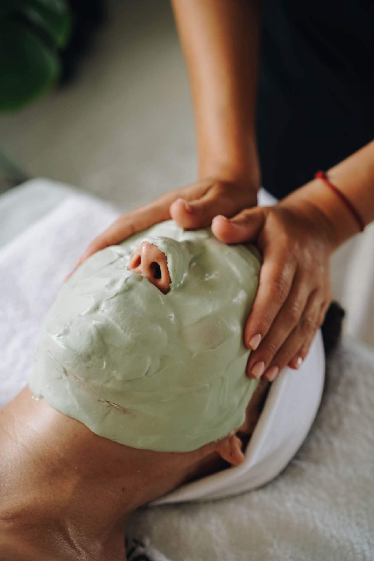 Hai bàn tay ôm nhẹ khuôn mặt phủ mặt nạ đất sét xanh của người nằm trên khăn bông, nhìn chéo từ trên