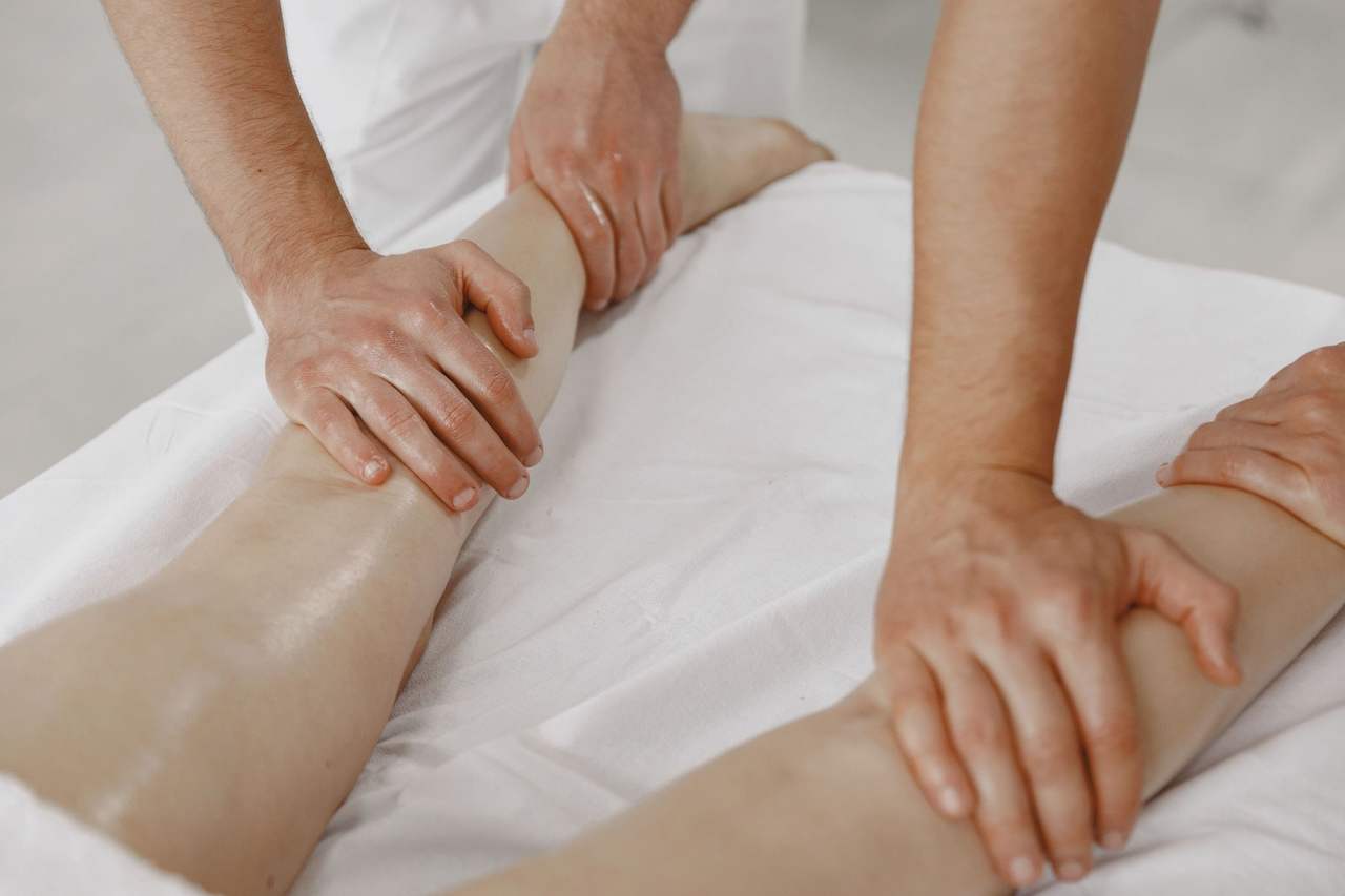 Bàn tay hai kỹ thuật viên nam cùng xoa bóp bắp chân một người nằm trên ga trắng