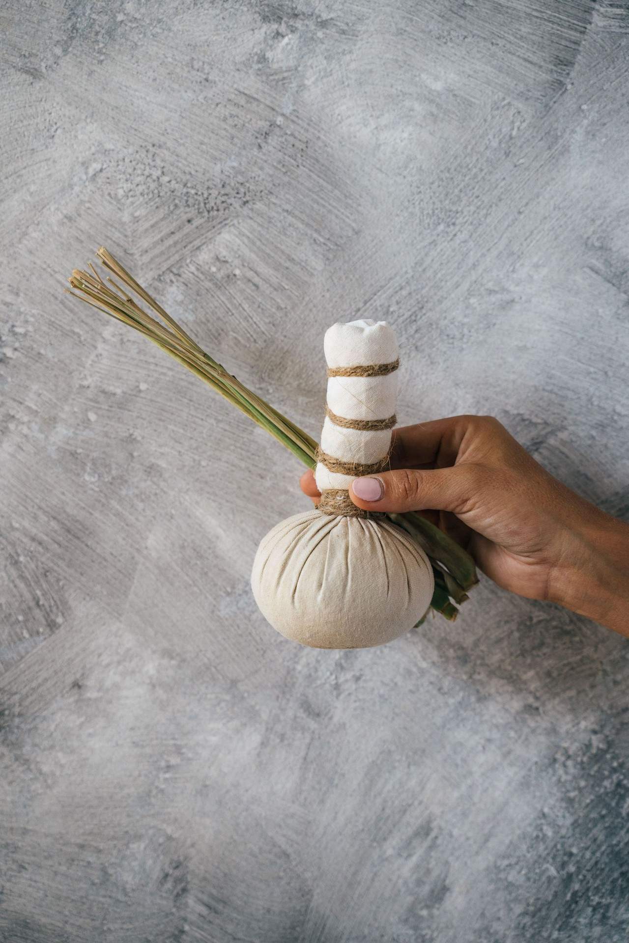 Một bàn tay cầm túi chườm thảo mộc bọc vải trắng buộc dây, kèm vài cọng sả, trên nền xi măng xám nhạt