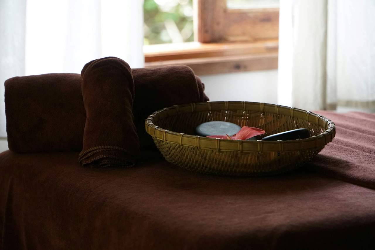 Giường trị liệu phủ khăn nâu bên cửa sổ gỗ, trên giường có khăn cuộn và chiếc rổ mây đựng đá cùng khăn nhỏ, ánh chiều qua rèm voan