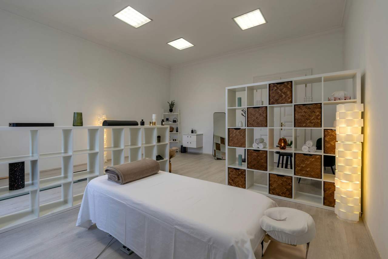 Phòng trị liệu tường trắng, sàn gỗ sáng, một giường massage phủ khăn trắng, kệ ô vuông có giỏ mây và khăn cuộn, đèn đứng tỏa ánh vàng nhẹ