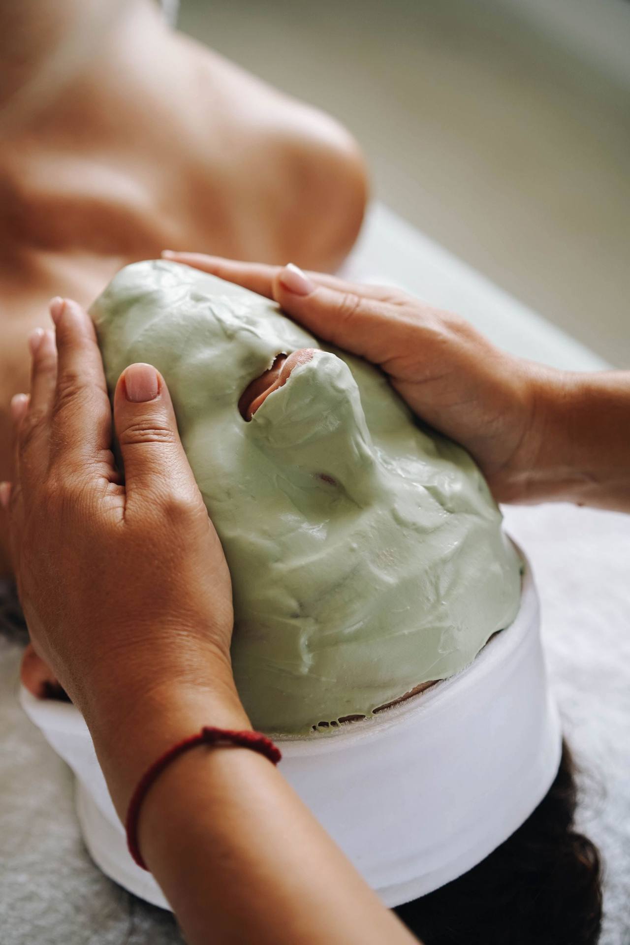 Hai bàn tay áp nhẹ hai bên khuôn mặt đã phủ kín mặt nạ đất sét xanh, băng đô trắng giữ tóc, cổ tay đeo sợi chỉ đỏ