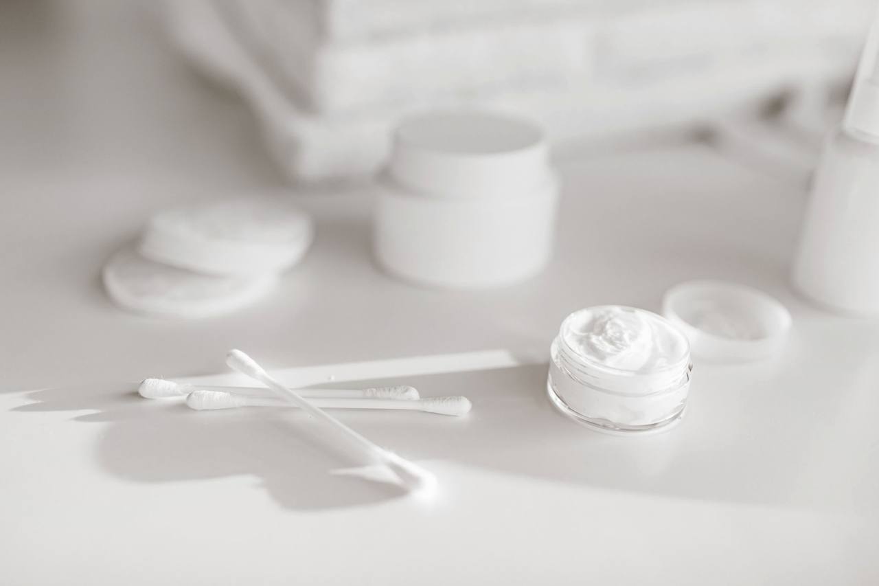 Trên mặt bàn trắng: vài miếng bông tẩy trang tròn, que bông, một hũ kem trắng mở nắp và lọ trơn không nhãn, khăn trắng mờ phía sau