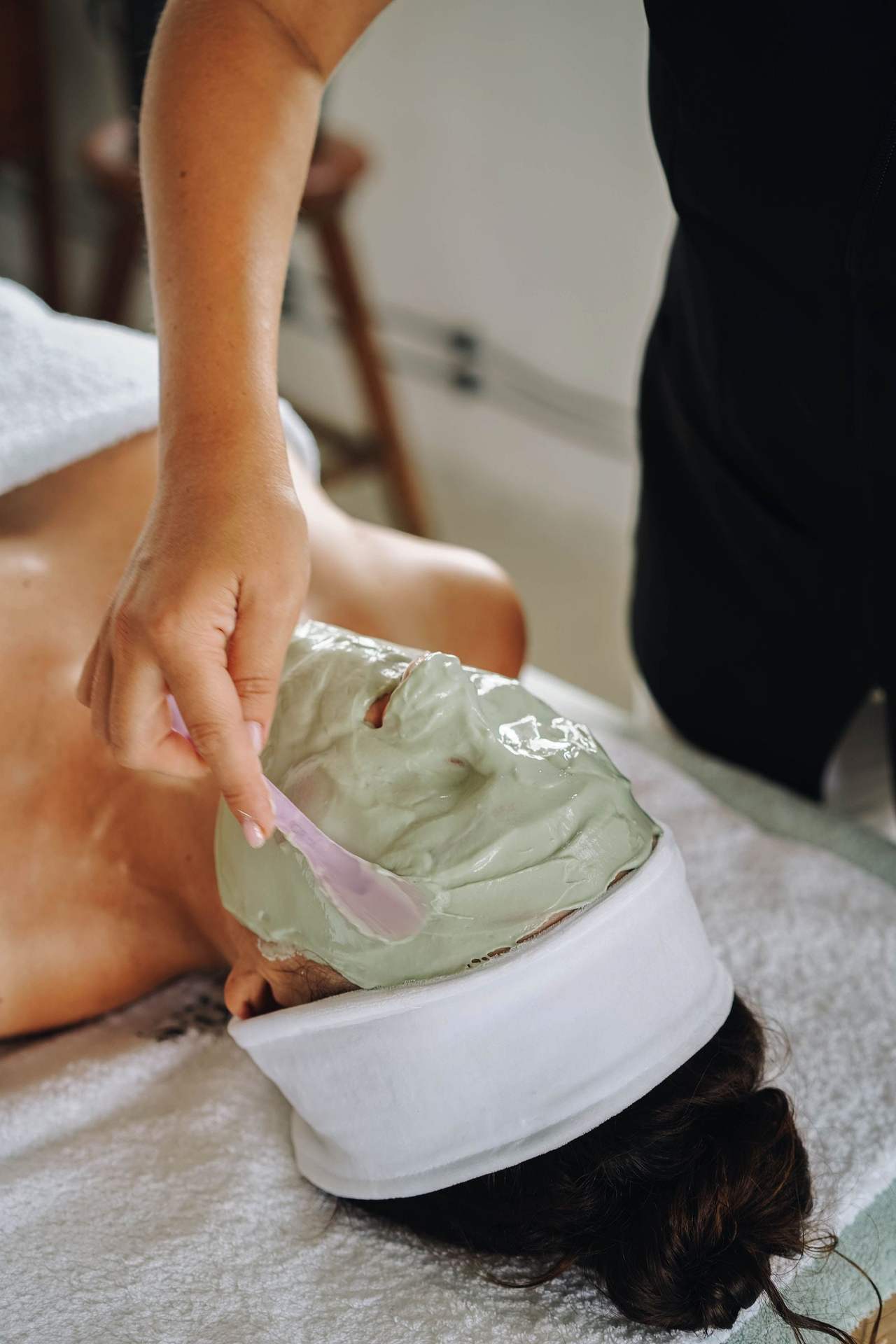 Bàn tay cầm cọ dẹt phết lớp mặt nạ đất sét xanh nhạt lên mặt một người đang nằm, tóc quấn băng đô trắng, đầu kê trên khăn bông