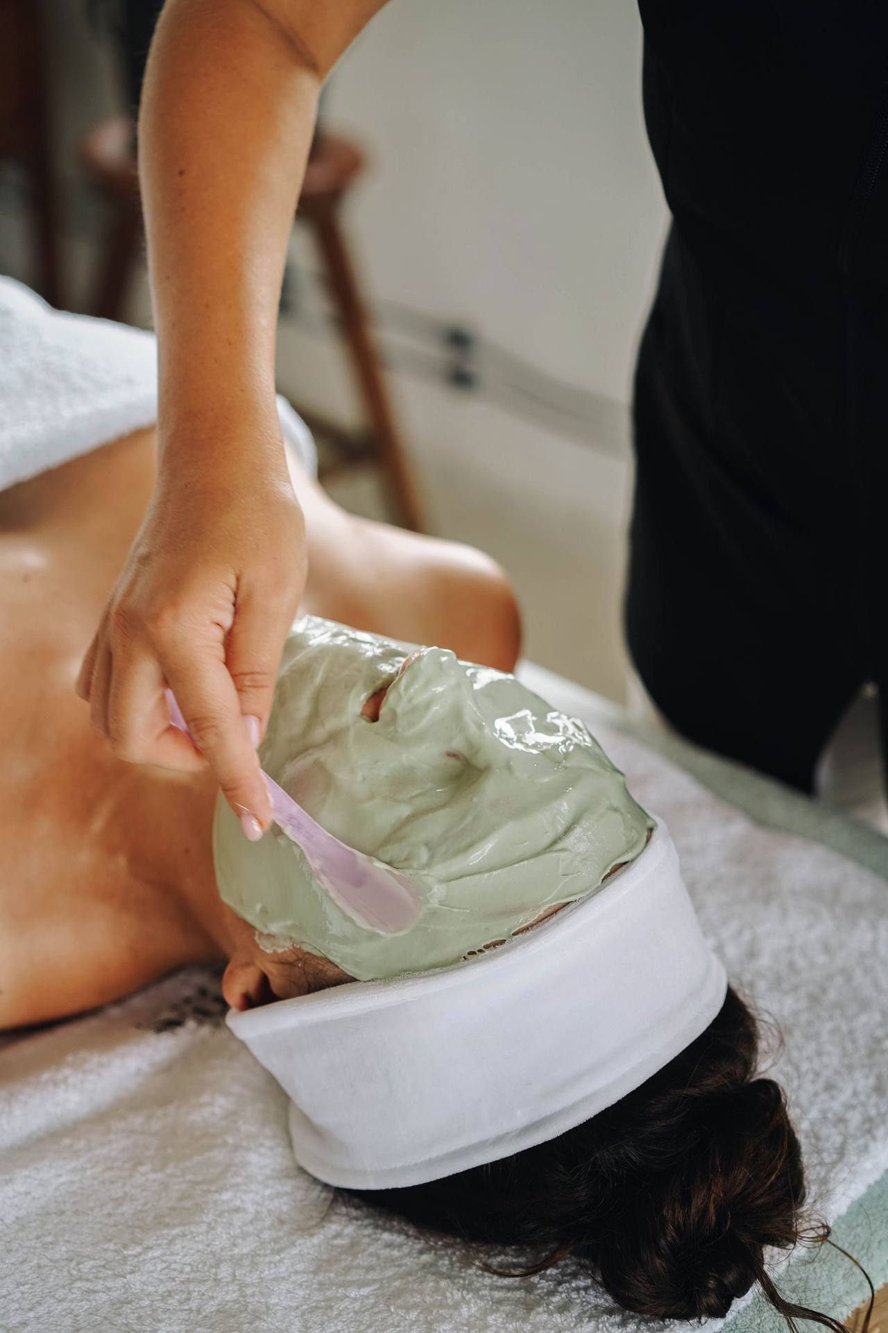 Bàn tay cầm cọ dẹt phết lớp mặt nạ đất sét xanh nhạt lên mặt một người đang nằm, tóc quấn băng đô trắng, đầu kê trên khăn bông