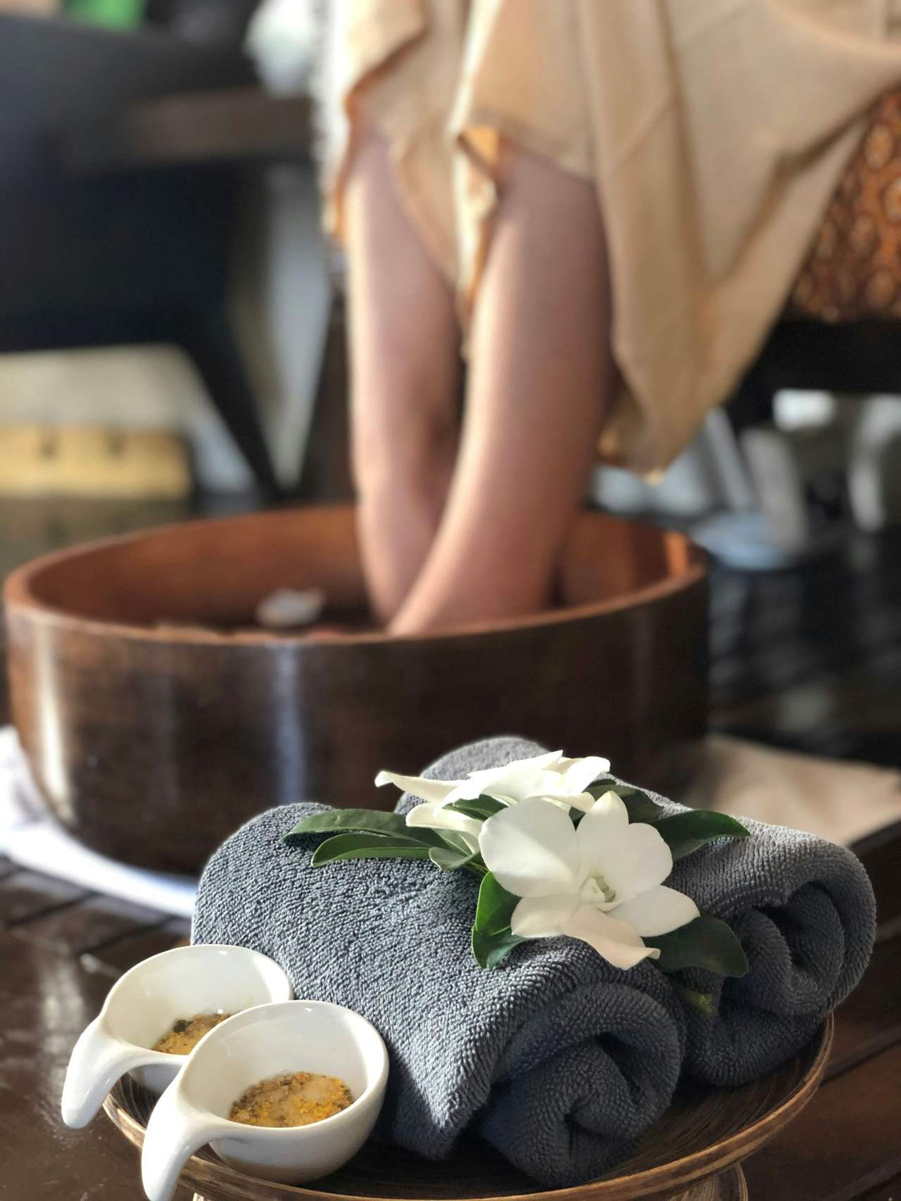 Đôi chân ngâm trong chậu gỗ sẫm màu; phía trước là khăn xám cuộn tròn cài hoa lan trắng và hai chén sứ nhỏ đựng muối trộn thảo mộc
