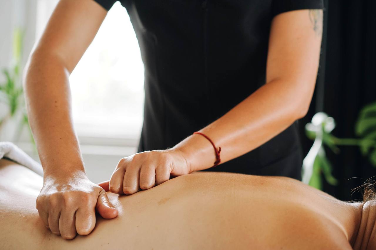 Hai bàn tay kỹ thuật viên áo đen ấn đều trên lưng trần của người nằm sấp, cửa sổ sáng và chậu cây phía sau