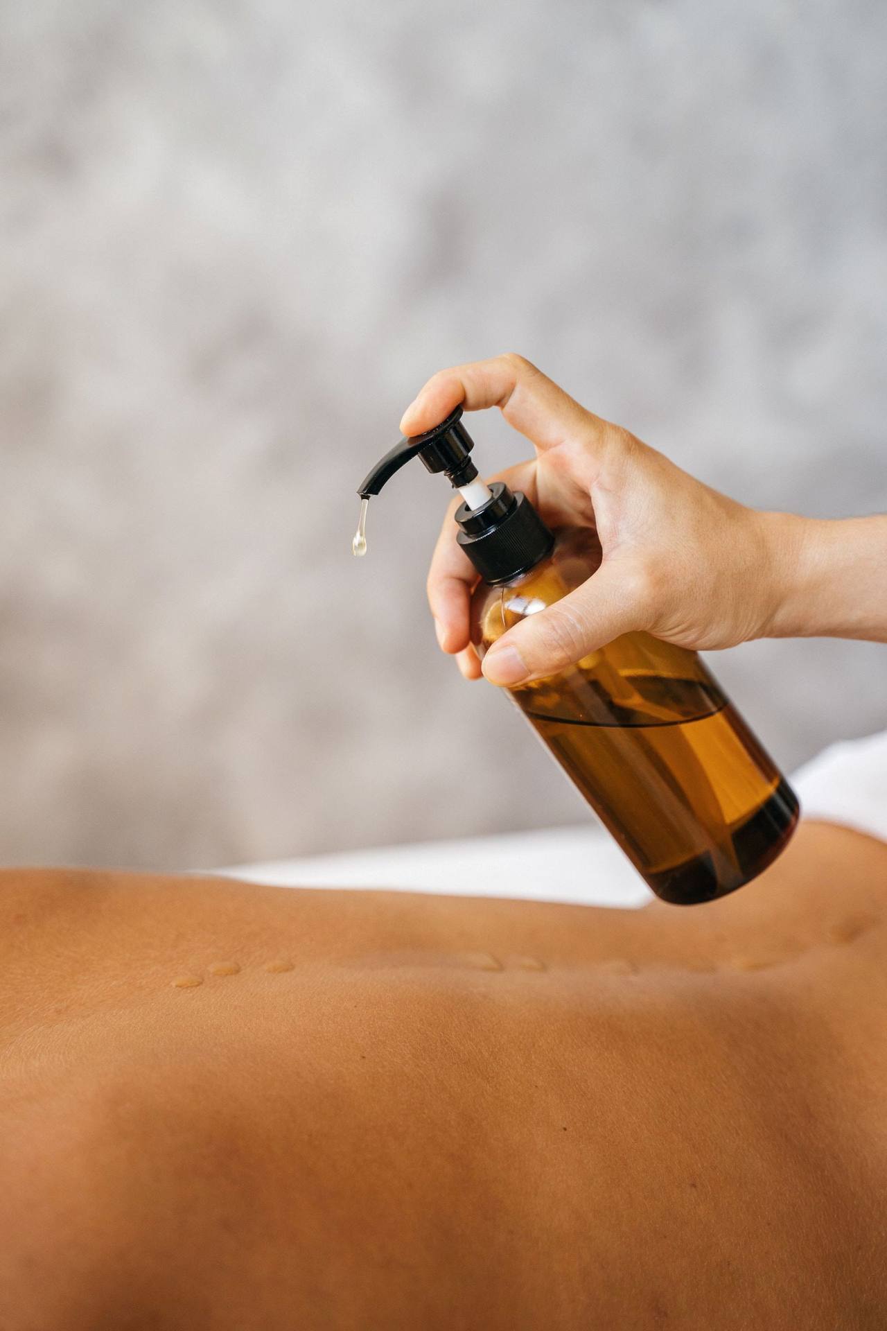 Một bàn tay bấm chai thủy tinh nâu có vòi, giọt dầu nhỏ xuống ngay trên lưng người đang nằm, tường xám nhạt phía sau