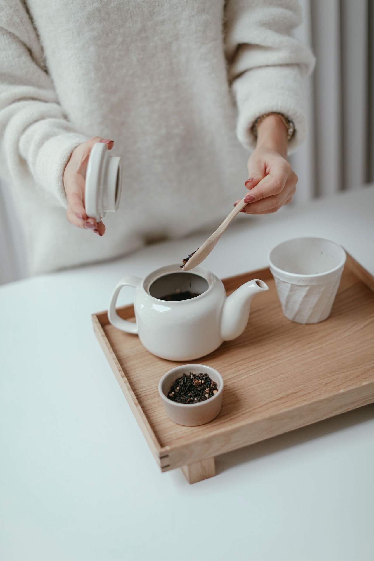 Hai bàn tay cho lá trà khô vào ấm sứ trắng trên khay gỗ sáng, bên cạnh chén trà và chén nhỏ đựng lá trà, mặt bàn trắng